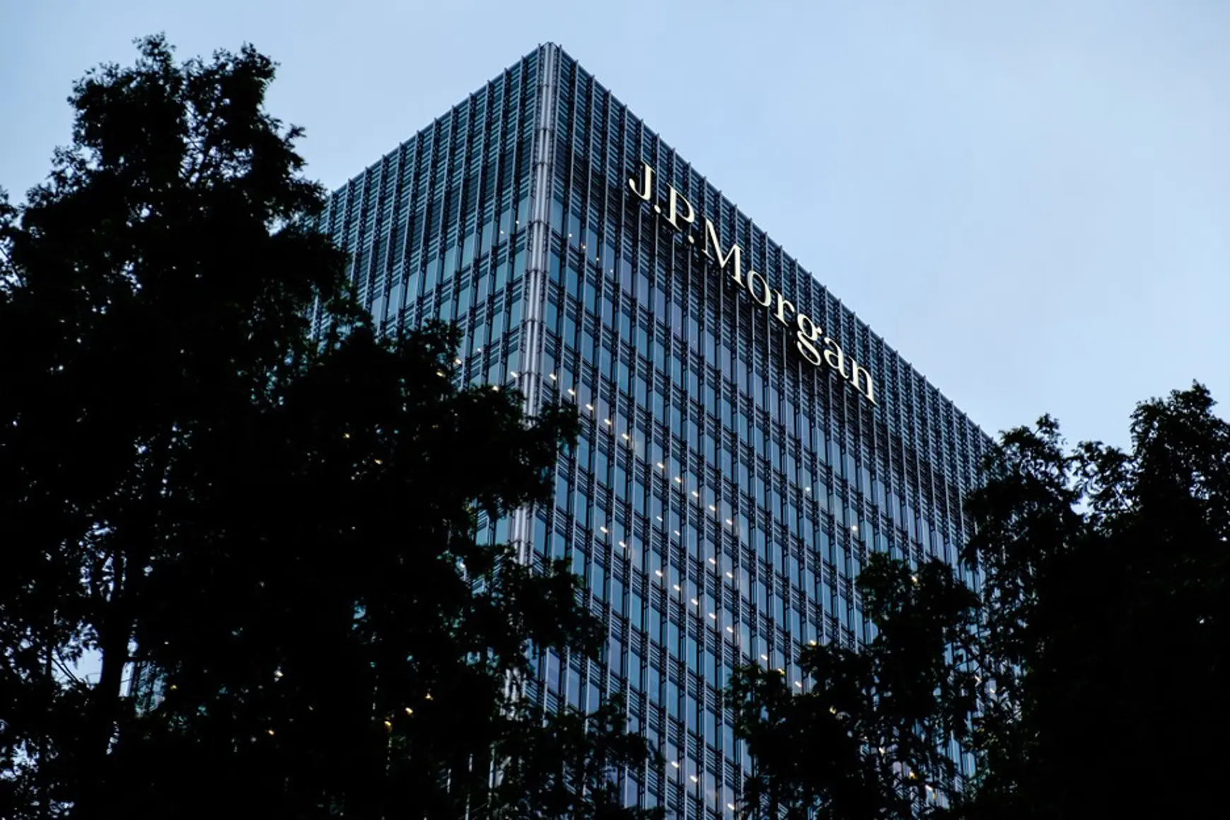 Een hoog glazen gebouw met het J.P. Morgan-logo erop staat tegen de achtergrond van bomen en lucht bij schemering. Het gebouw wordt van binnen verlicht door lampen die een sfeer van rust creëren.