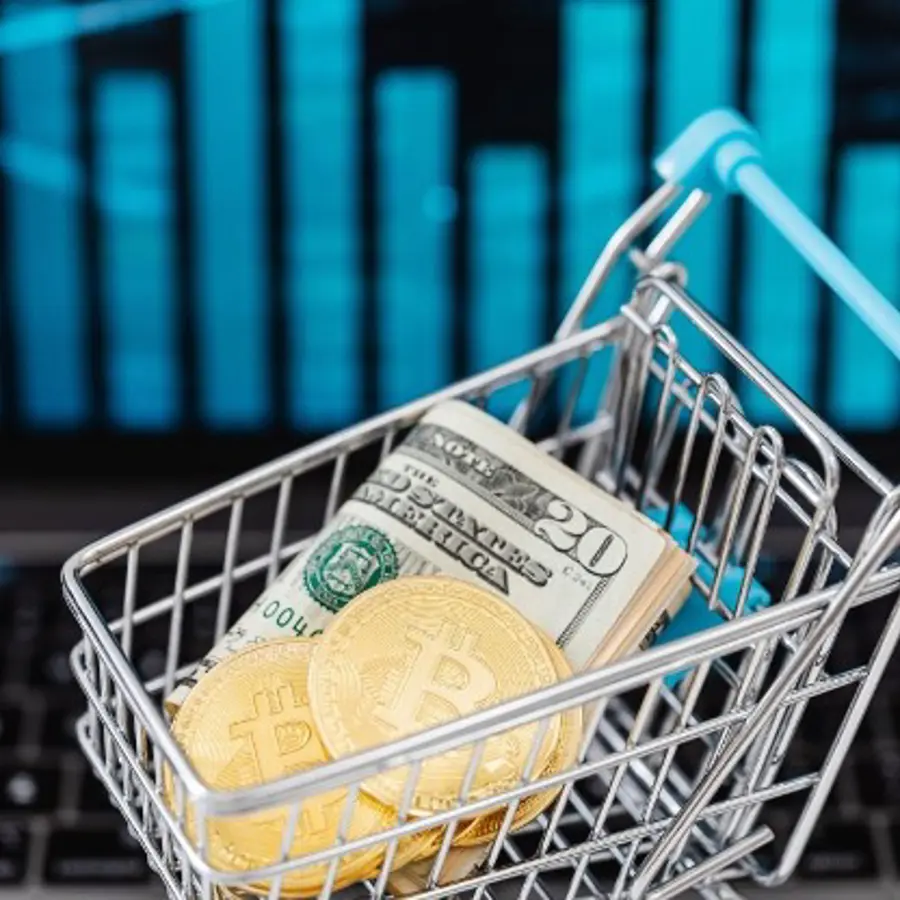 Winkelwagentje gevuld met Amerikaanse dollarbiljetten en Bitcoin-munten, op de achtergrond een laptop met grafiek.